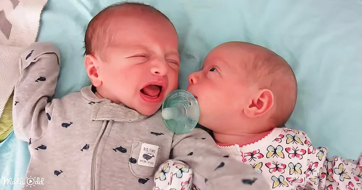 Two newborn babies, one yawning, the other holding a pacifier, lying on a light blue blanket.