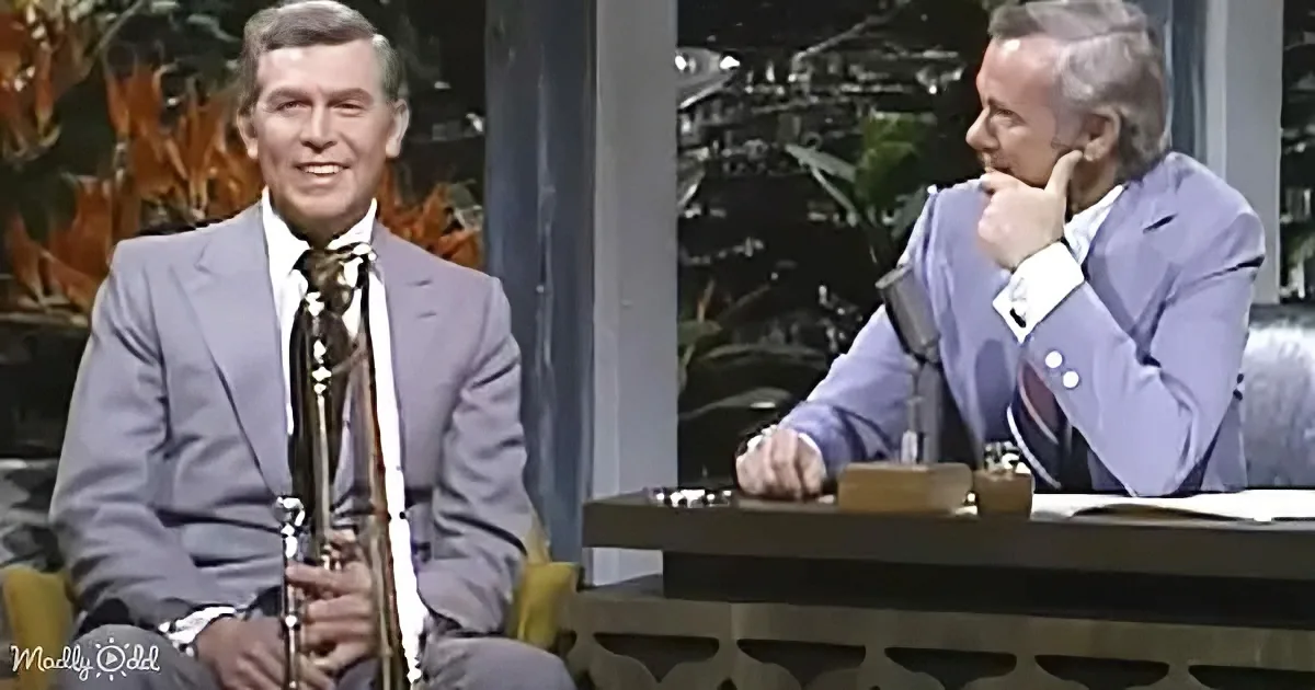 A man in a suit holds a clarinet, seated next to another man in a suit at a desk with a microphone.