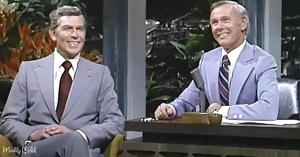 Two men in suits smile on a talk show set with a desk and microphone.