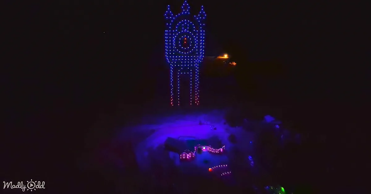 A towering, illuminated structure resembling a clock or robot stands atop a hill, glowing in blue and red against a dark sky.