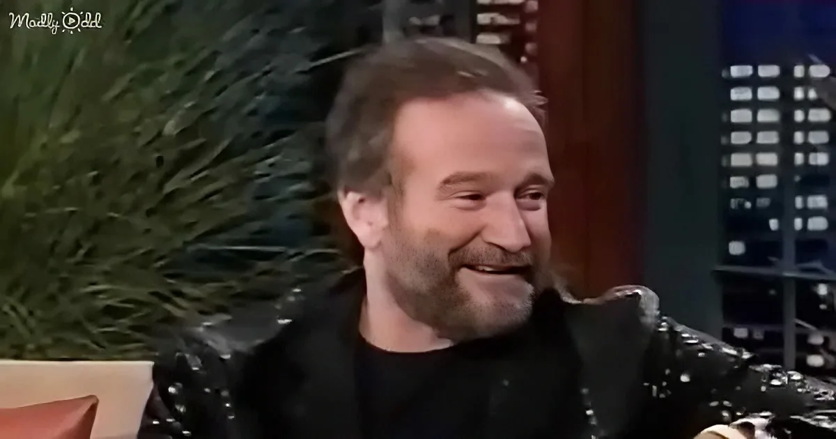 A man with a beard and short hair is smiling while sitting on a couch, wearing a black jacket and black shirt.
