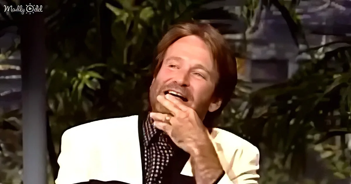 A man in a white suit jacket and checkered shirt is laughing with his hand on his mouth.