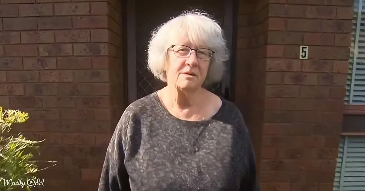 An elderly woman with white hair and glasses stands in front of a brick building with the number 5 visible.