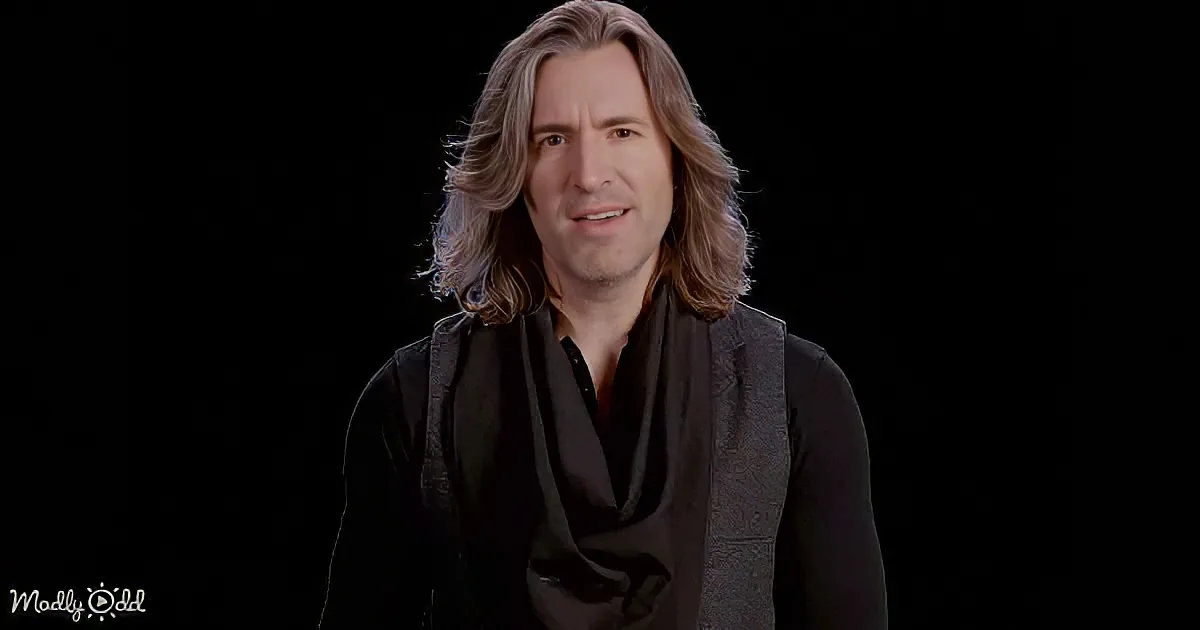 A man with long, wavy hair and a black outfit stands against a dark background.