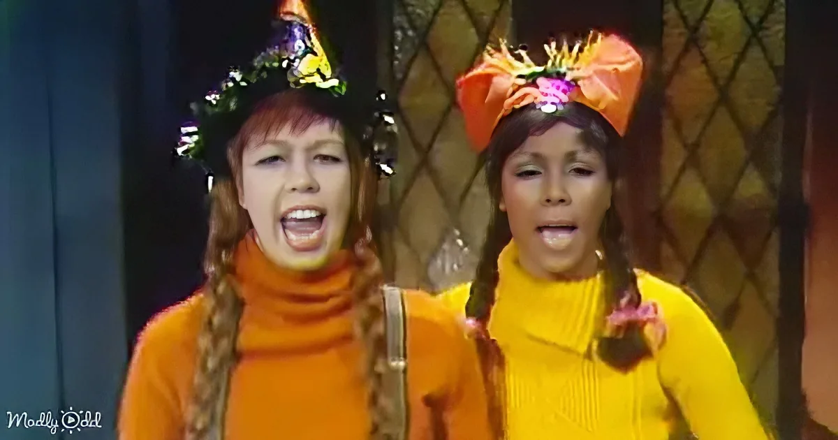 Two individuals in vibrant orange outfits with floral headpieces, singing or speaking with expressive faces.