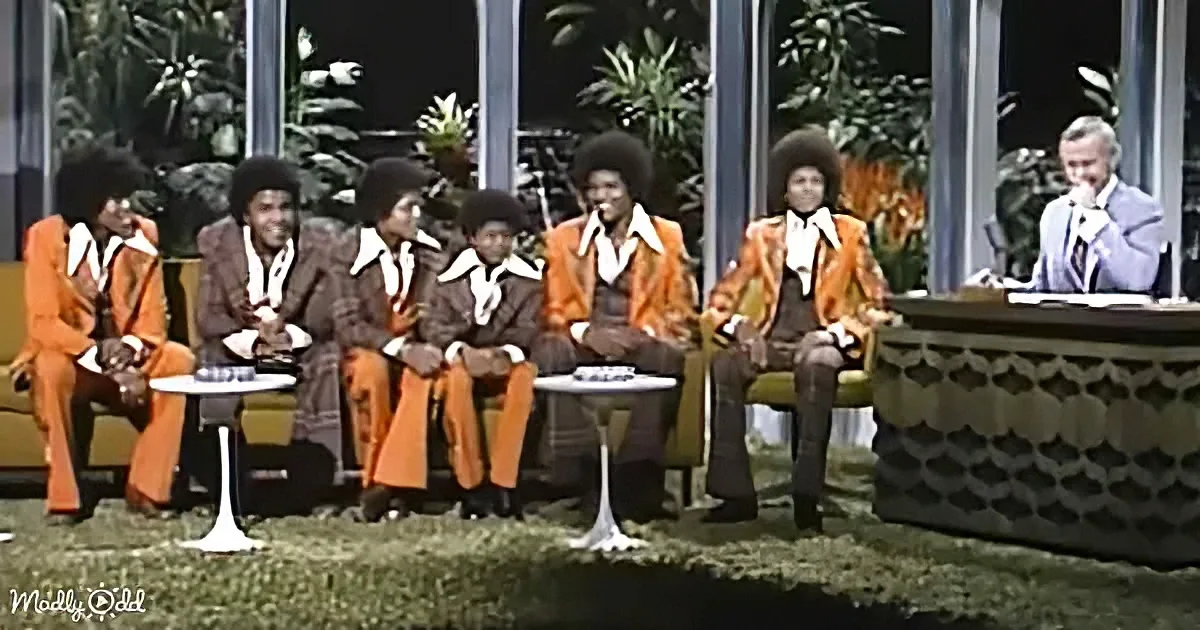 The Jackson 5, dressed in vibrant suits, sit on a stage with a host, surrounded by lush greenery and modern decor.