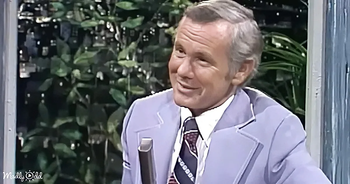 A man in a light purple suit and tie smiles while holding a microphone, with greenery in the background.