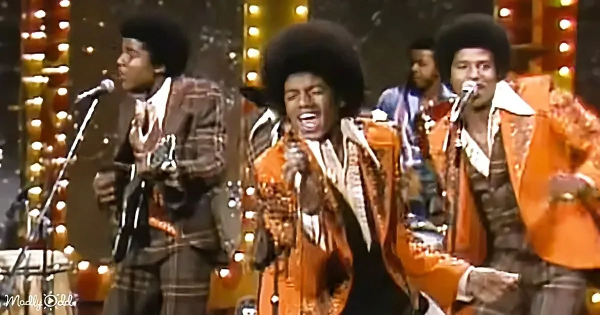 A man in a vibrant orange suit sings passionately into a microphone on stage.