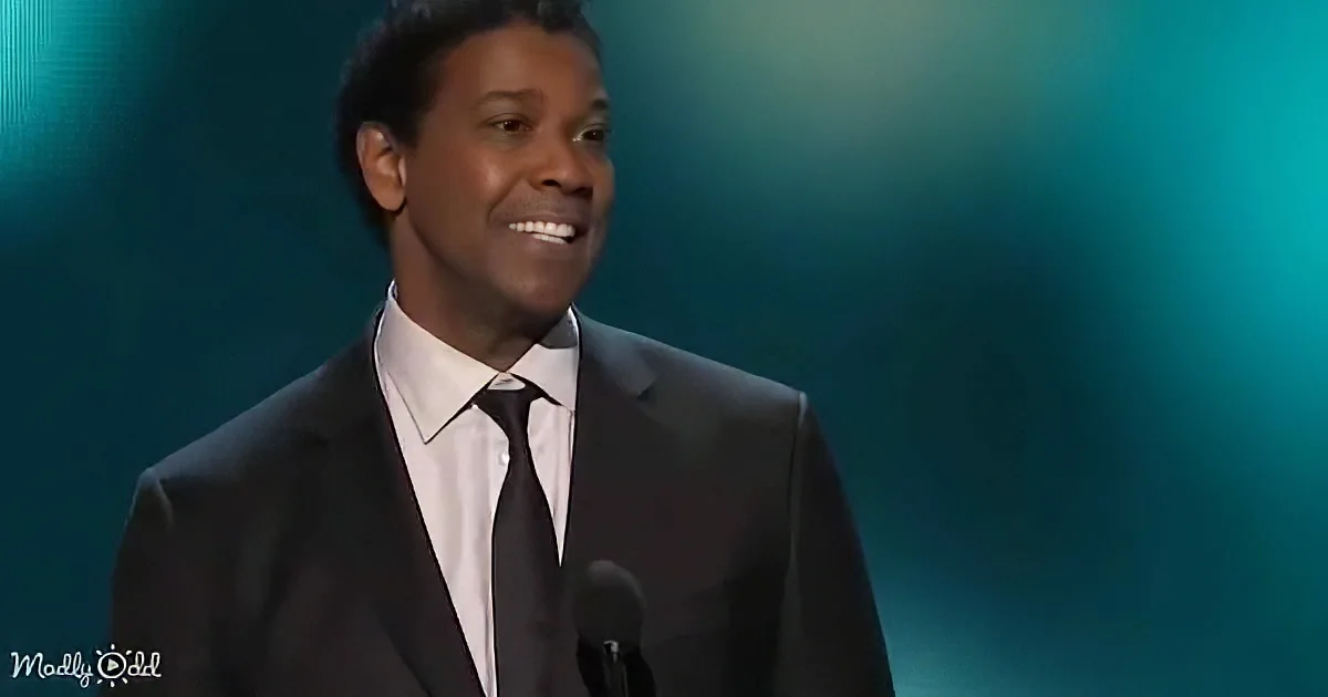 A man in a black suit and white shirt smiles at a podium with a microphone, against a teal background.
