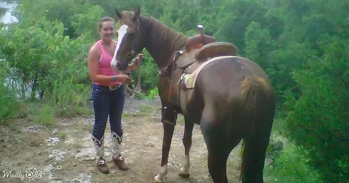 A woman in pink tank top and jeans stands beside a saddled horse, holding its reins, with lush greenery in the background.