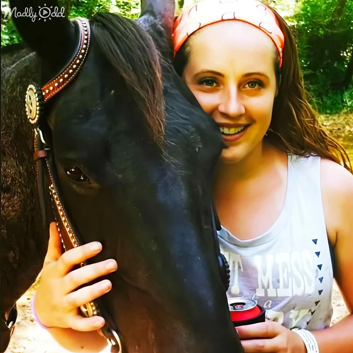 A woman smiles closely with a black horse, wearing a headband and tank top, in a natural setting.