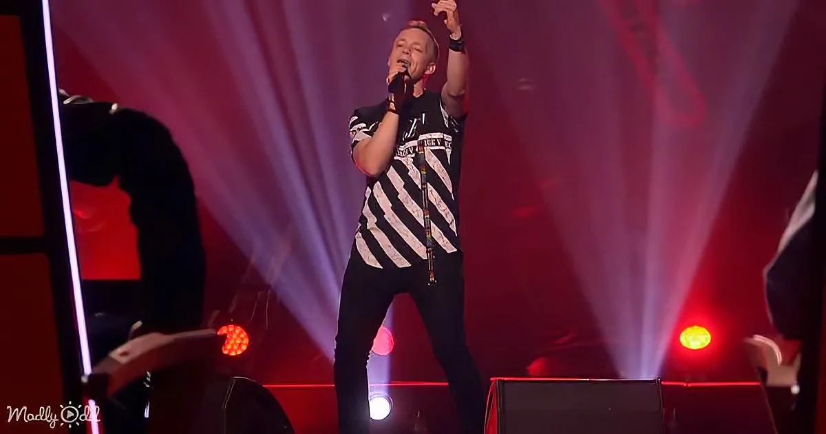 A man in a black and white striped shirt sings on stage with red and white lights.