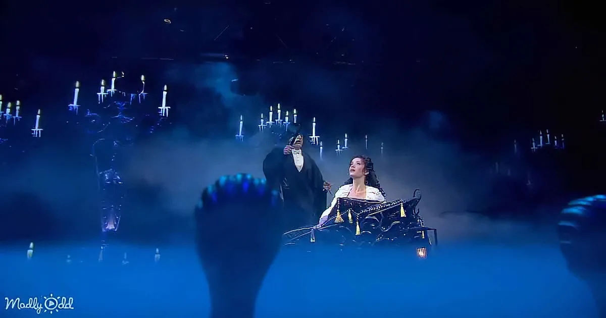 A man in a black robe sings passionately as a woman sits in a carriage, surrounded by candles and fog.