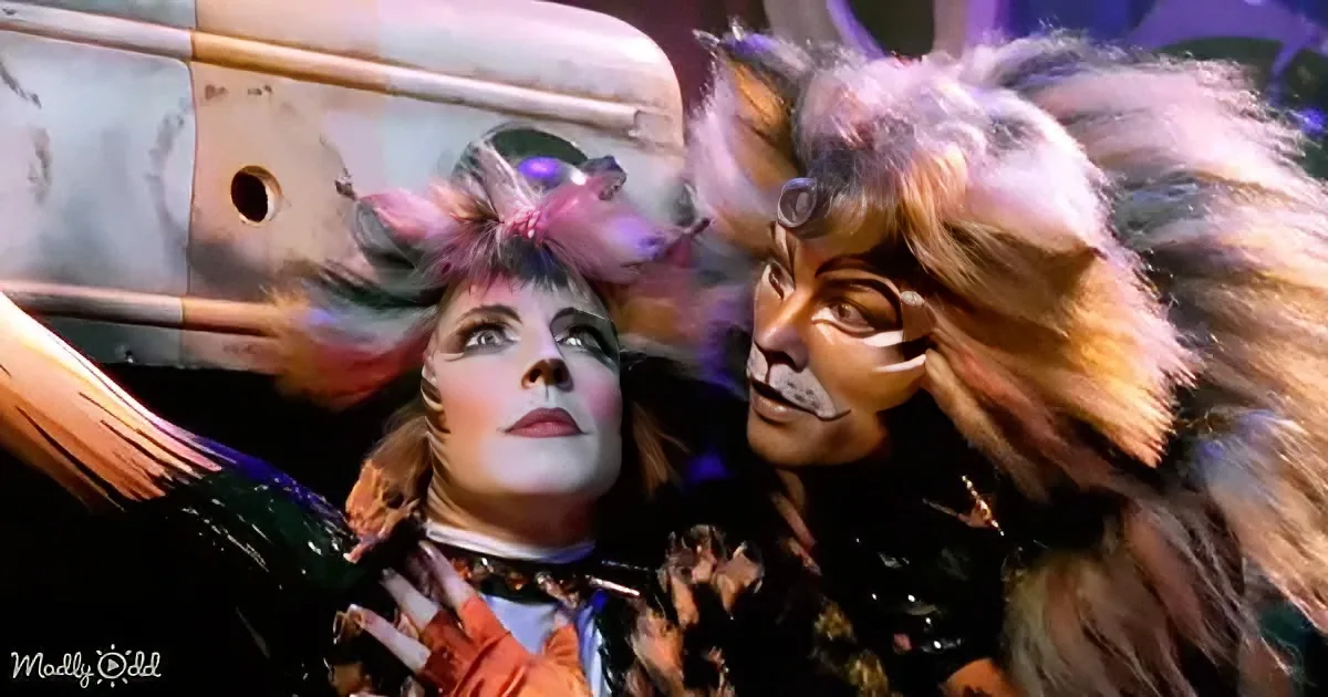 A performer in a colorful, elaborate costume with cat-like features is being embraced by another performer in a similar...