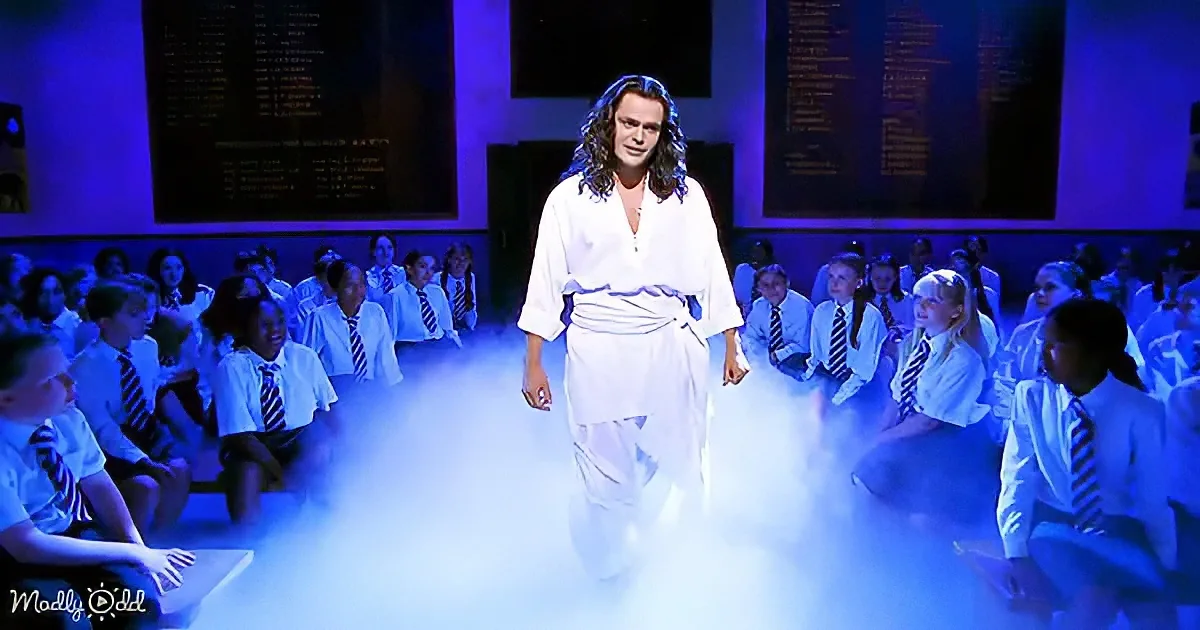 A person in white robes stands on a stage surrounded by seated students in school uniforms.