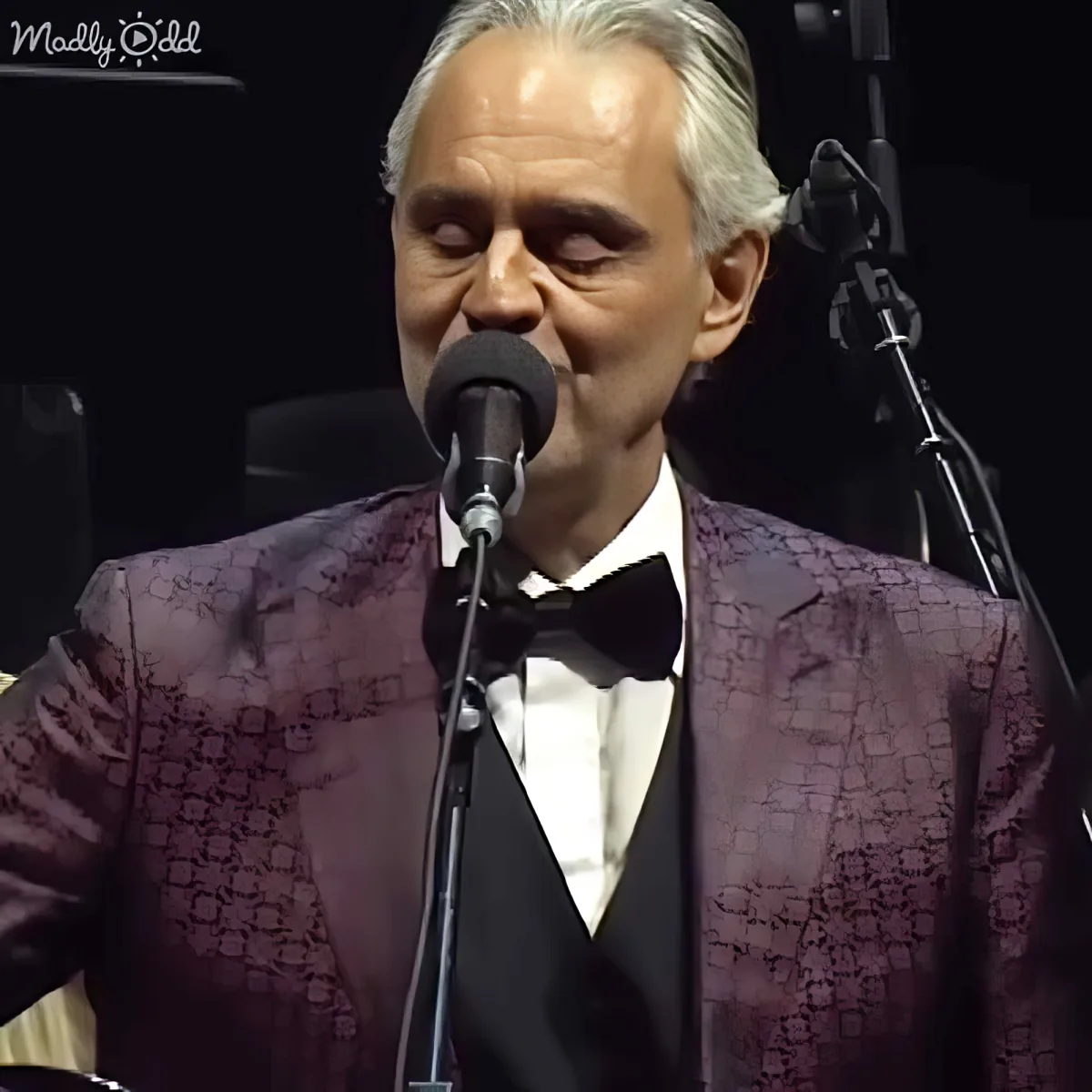 A man in a purple patterned suit and bow tie sings into a microphone on stage.