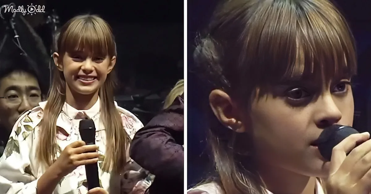 A young girl with long hair and bangs holds a microphone, smiling and singing on stage.
