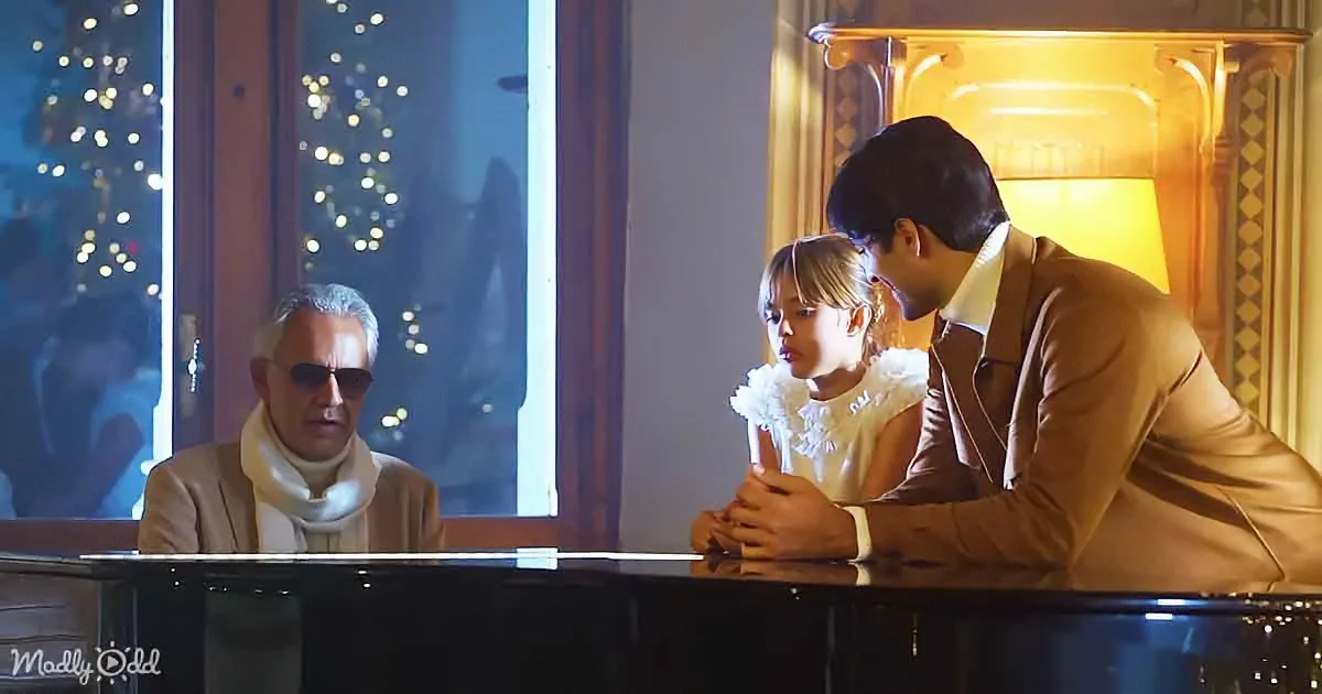 A man in a brown suit leans over a piano, a young girl in white sits beside him, both focused on the instrument.