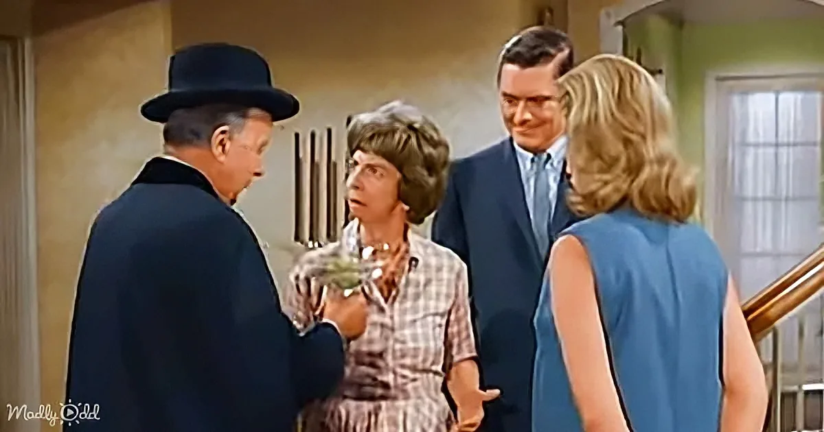 A man in a dark suit and hat is holding a glass object, interacting with a woman in a plaid dress and another woman in a...