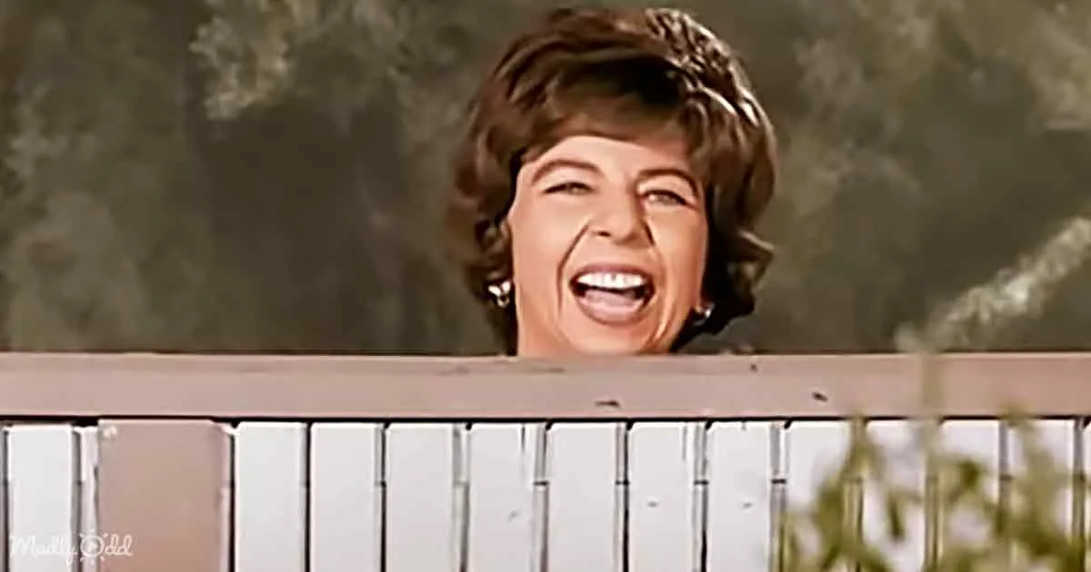 A woman with short brown hair and a wide smile is peeking over a white fence.