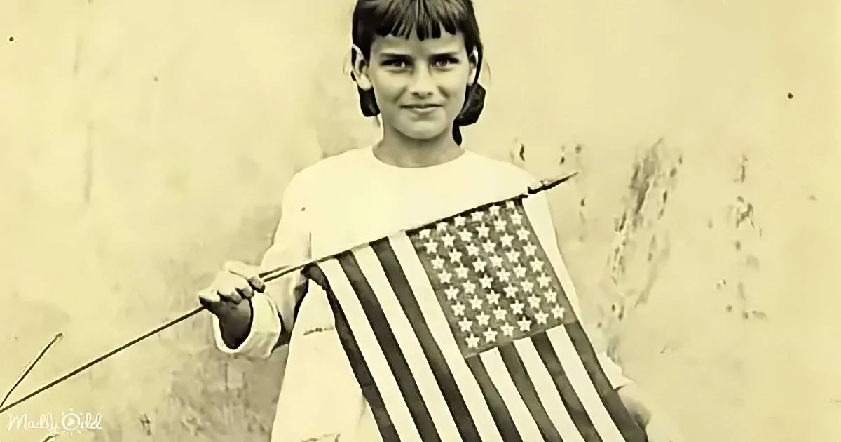 A young girl holds a small American flag, smiling, in a vintage black-and-white photo.