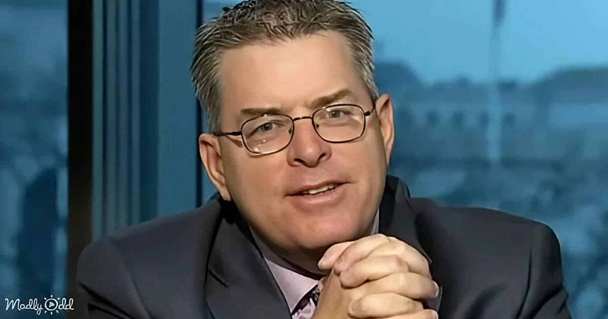 A man in a suit and glasses sits with clasped hands, looking slightly to the side.