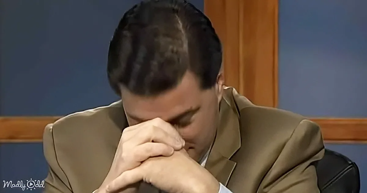 A man in a suit sits with his head down, hands clasped, appearing contemplative or distressed.