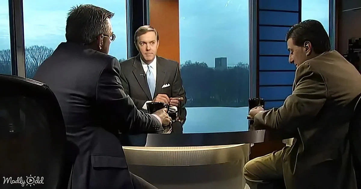 A man in a suit and tie is seated at a table with two others, discussing in a studio setting.