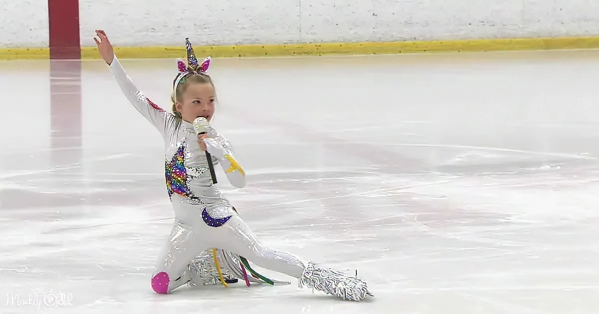 A child in a silver sequined costume with a unicorn headband sings into a microphone on an ice rink.