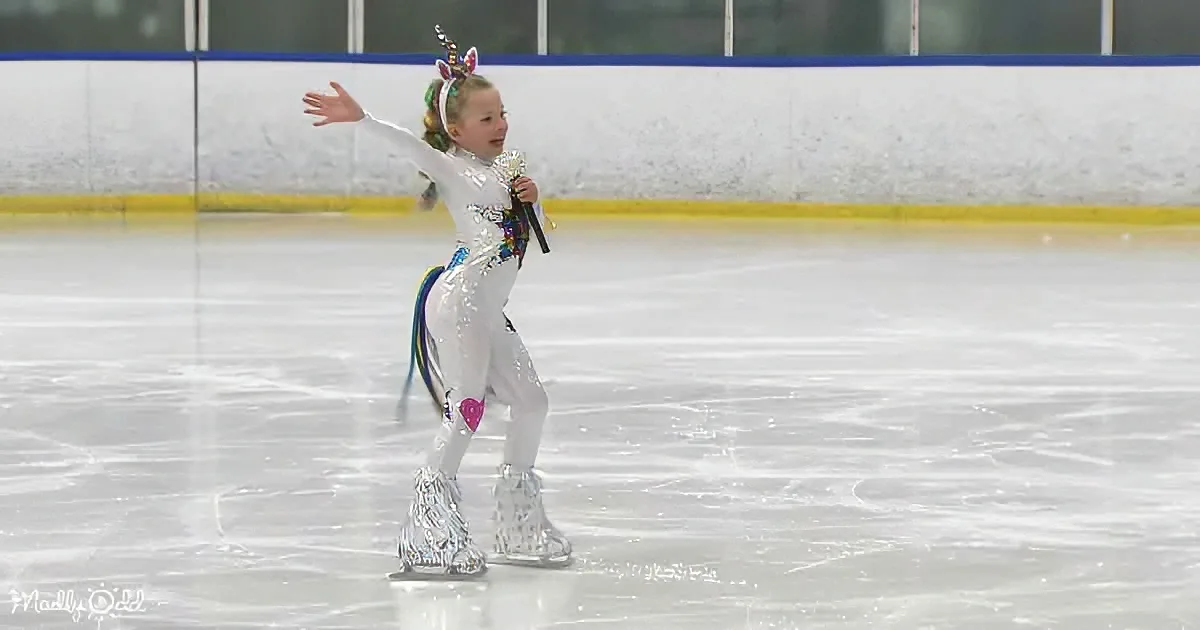 A child in a white, sparkly unicorn costume skates on an ice rink, holding a microphone.
