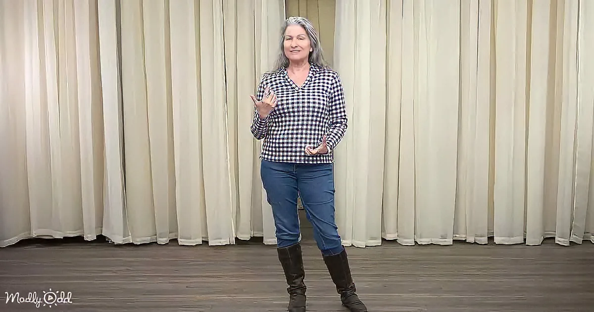 A woman in a checkered shirt and jeans stands in front of a curtain, gesturing with her hands.