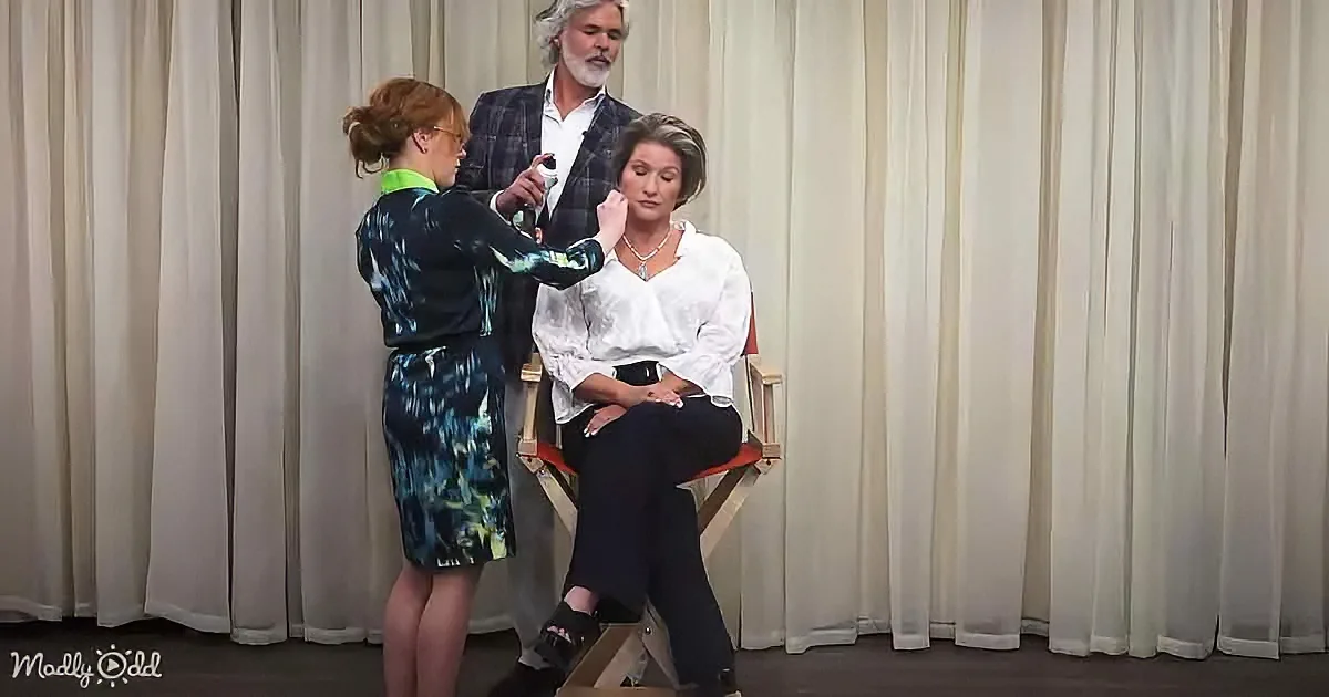 A woman in a floral dress applies makeup to another woman seated in a director's chair.