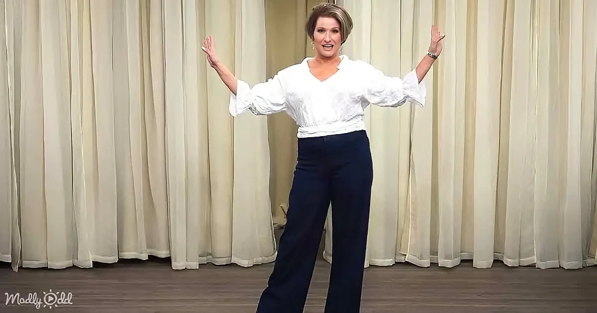 A woman in a white blouse and dark pants stands with arms raised, smiling, in front of a cream curtain backdrop.