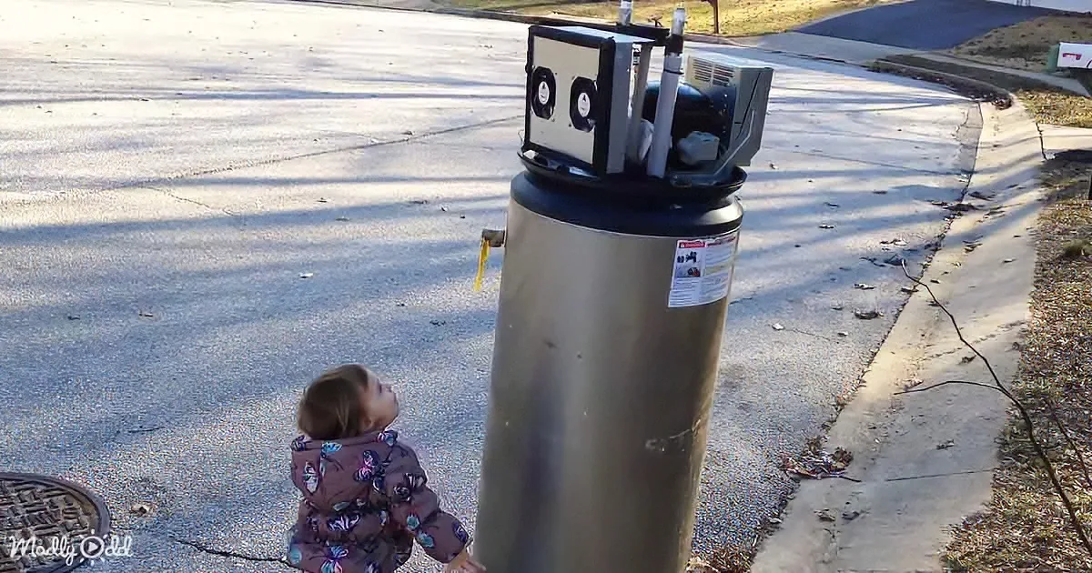 A child in a floral jacket stands beside a large, metallic water heater on a sunny day.