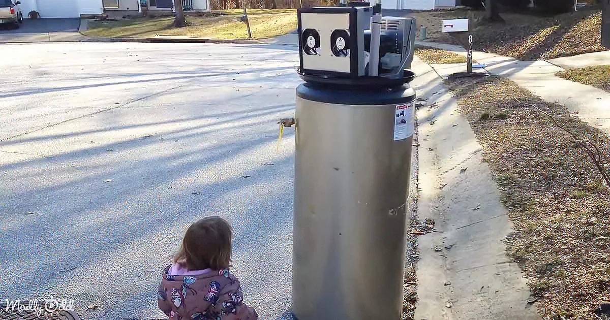 A child in a floral jacket stands near a large water heater on a residential street.