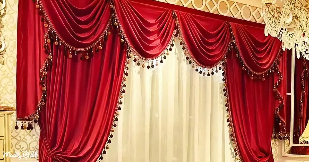 A luxurious red curtain with gold tassels hangs elegantly in a room with ornate wallpaper and a chandelier.