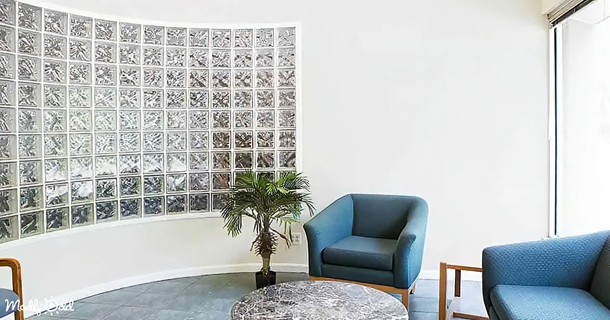 A modern room features a glass block wall, blue armchairs, a potted plant, and a marble coffee table.