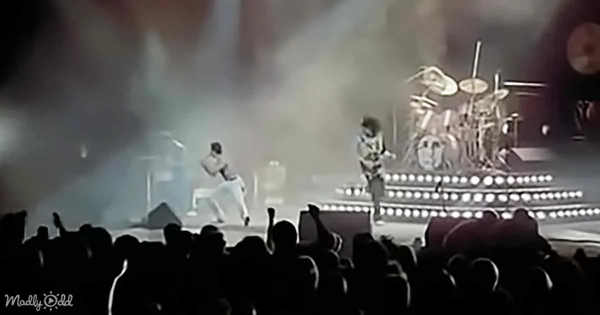 A performer in white pants and a black top dances on stage with a drummer behind, surrounded by bright lights and a crowd...