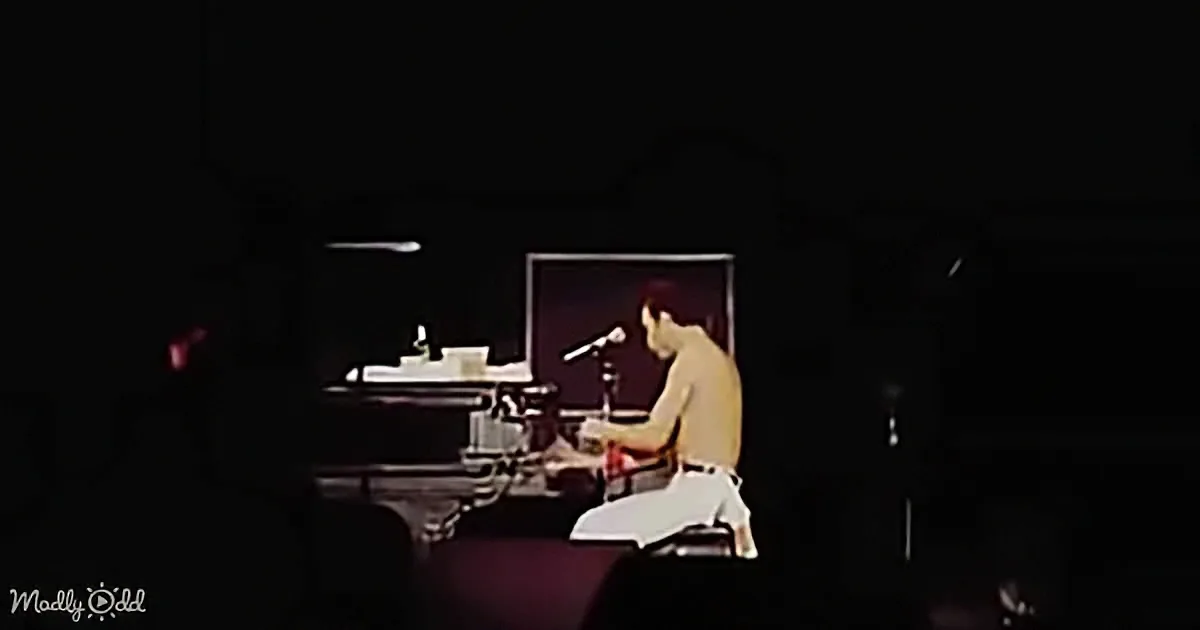 A person sits at a piano on stage, illuminated by a spotlight, with a microphone in front of them.