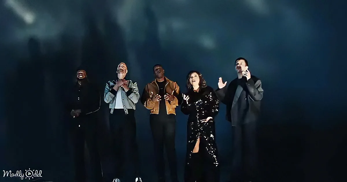 A group of five people, including a woman in a sequined dress, stand on stage, clapping and smiling under dim lighting.