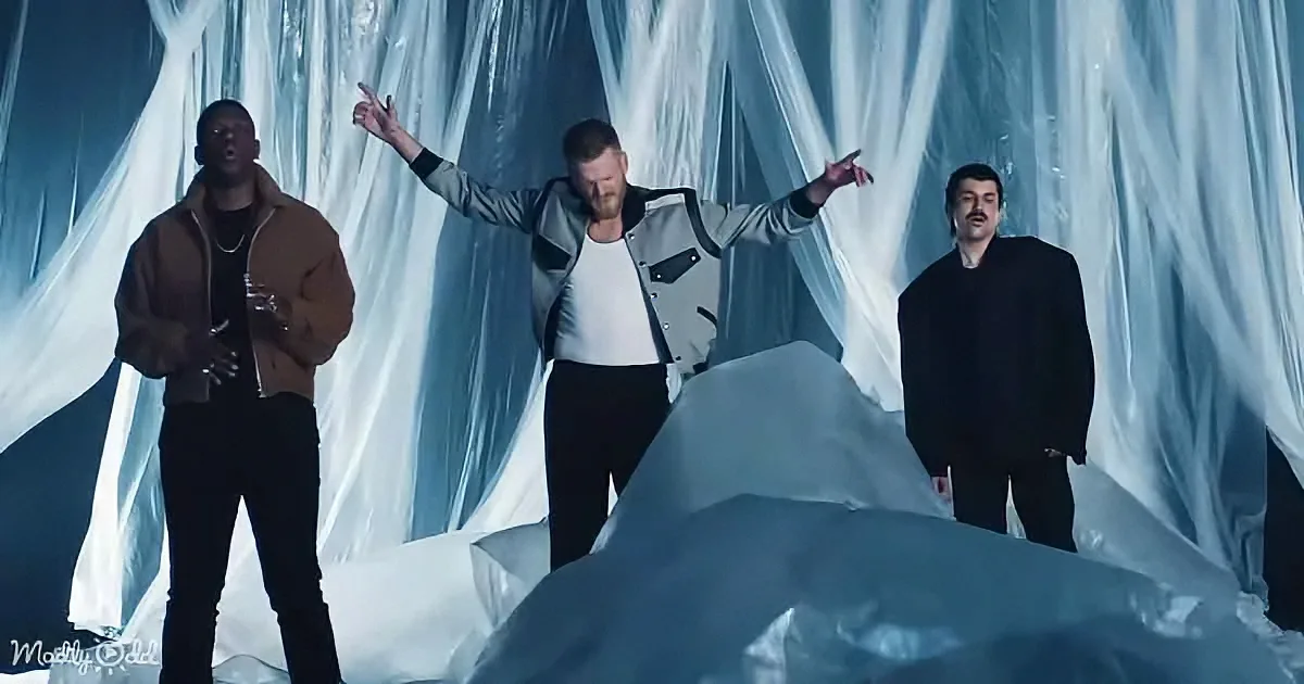 A man in a silver jacket stands with arms outstretched, surrounded by draped fabric, with two others nearby.