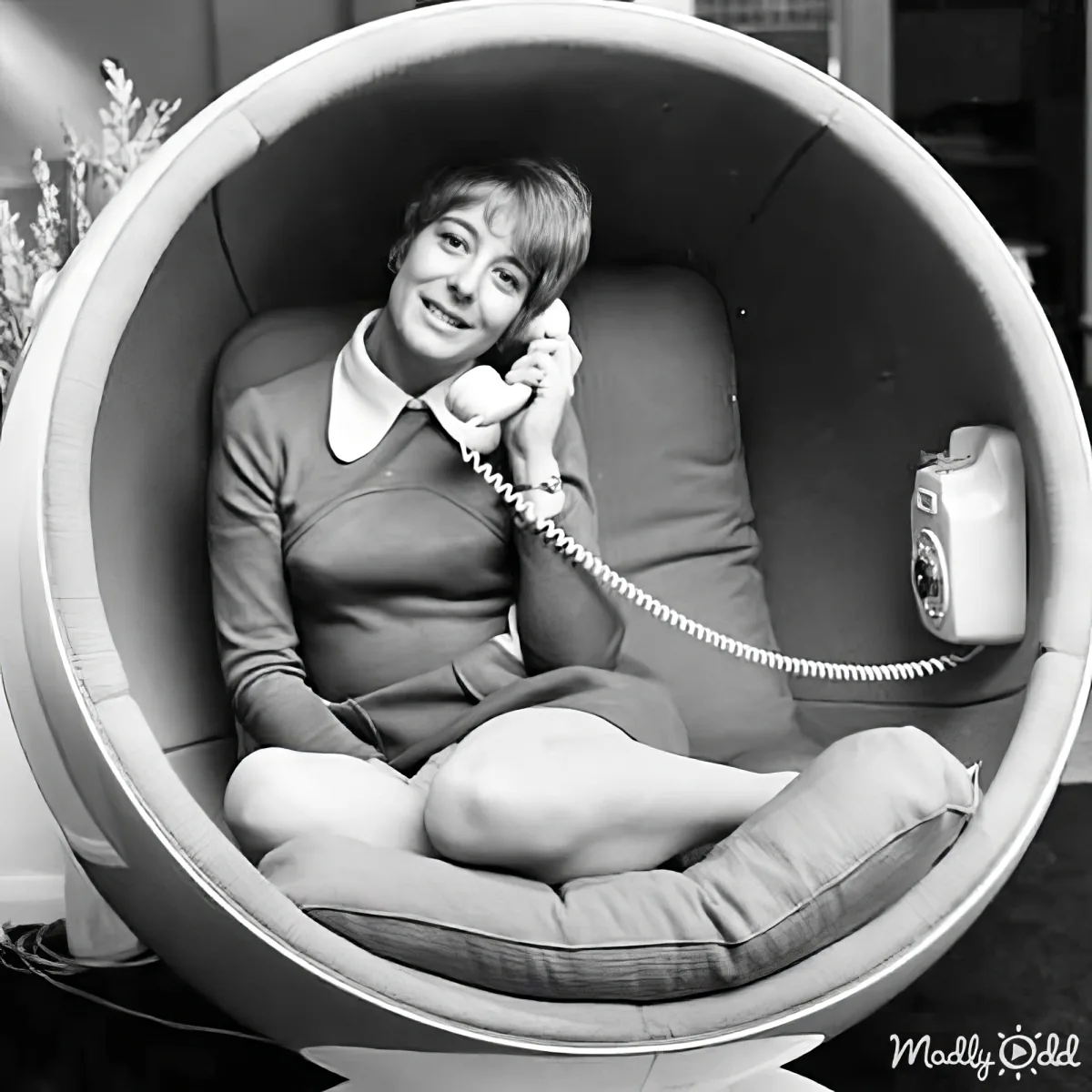 A woman sits in a futuristic, round, cushioned pod chair, holding a telephone receiver.
