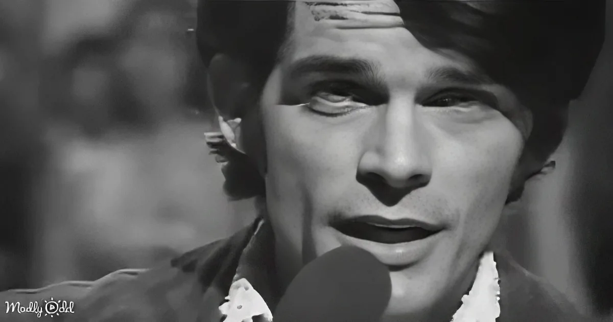 A close-up of a person singing into a microphone, wearing a patterned shirt, with a blurred background.