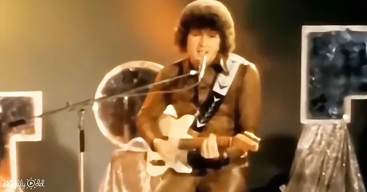 A person in a brown outfit plays a white guitar on stage with a microphone.