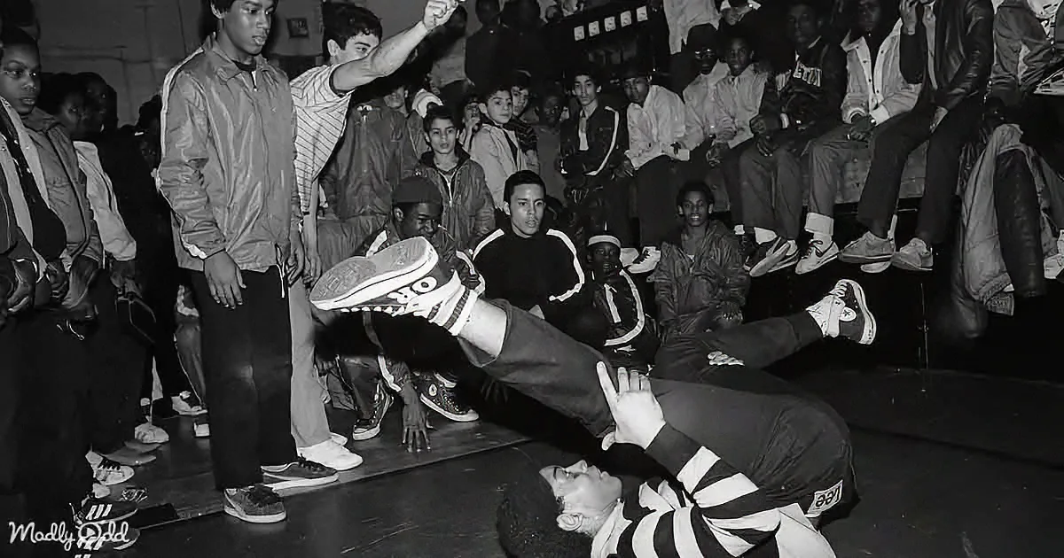 A dancer in a striped shirt performs a breakdance move on the floor, surrounded by an audience watching.