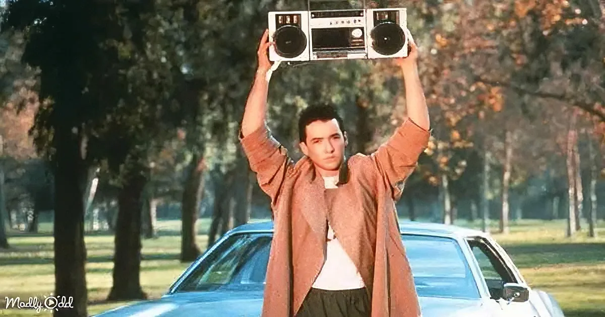 A man holds a boombox above his head, standing in front of a car in a park-like setting.