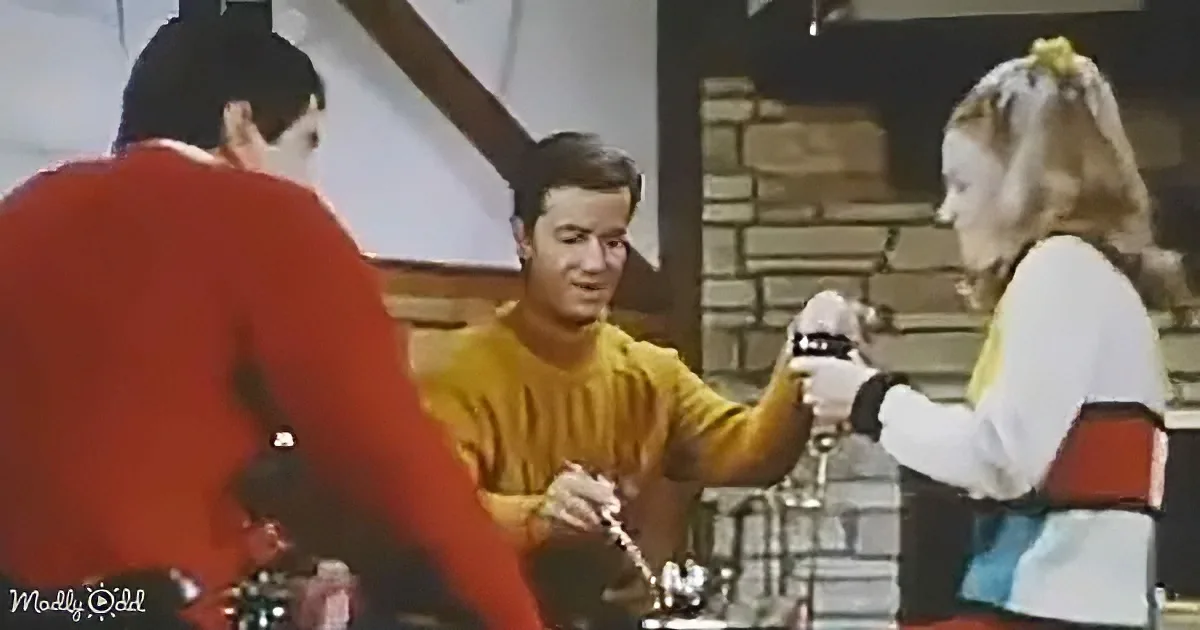 A man in a yellow shirt holds a device, interacting with a woman in a white and red outfit, while another person in red...