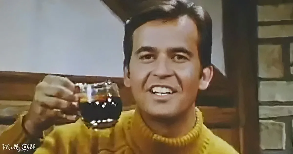 A man in a yellow sweater holds a glass of dark liquid, smiling, with a wooden structure and stone wall in the background.
