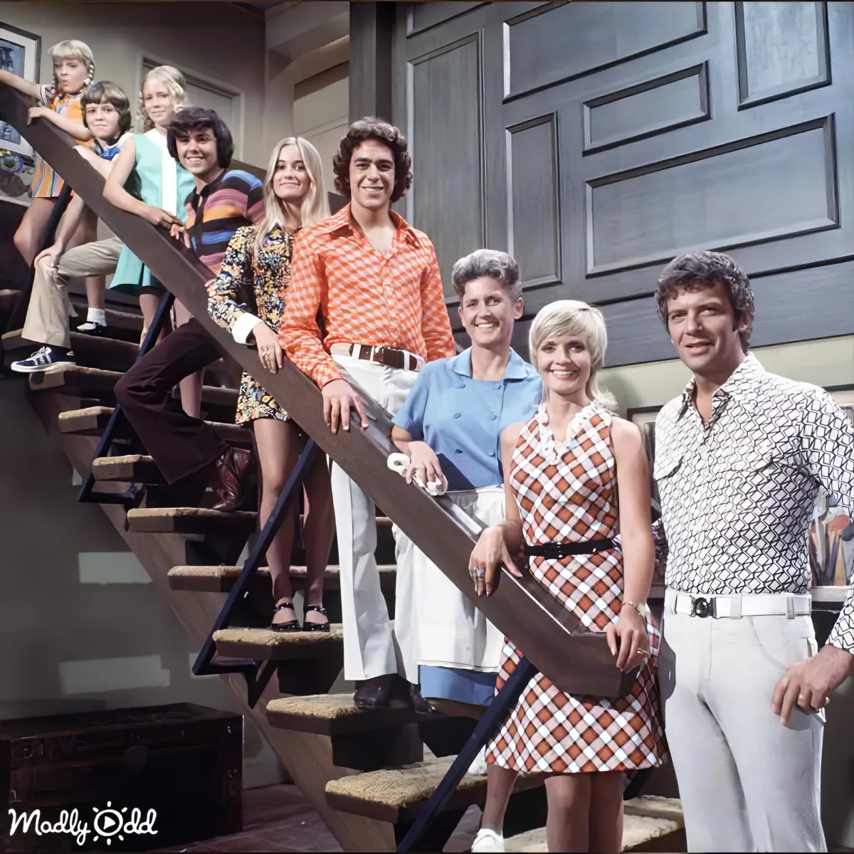 A family of nine stands on and around a staircase, dressed in 1970s fashion, smiling and posing together.