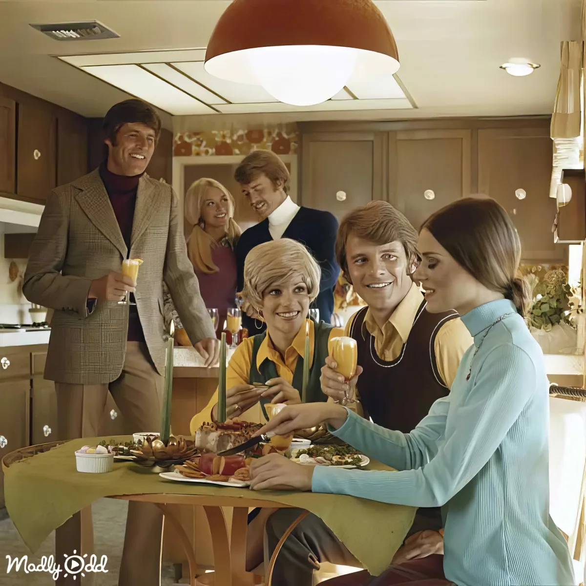 A group of people in a 1970s kitchen, dressed in vintage attire, gather around a table with food and drinks, smiling and...